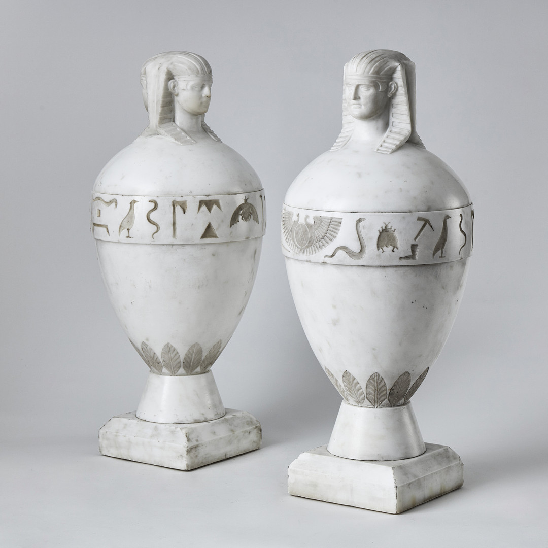 KANOPER RETOUR d'EGYPTE, ett par, Italien 1800-talets första hälft, skulpterad carraramarmor, krönta av dubbelsidiga Antinous-Osiris huvuden.