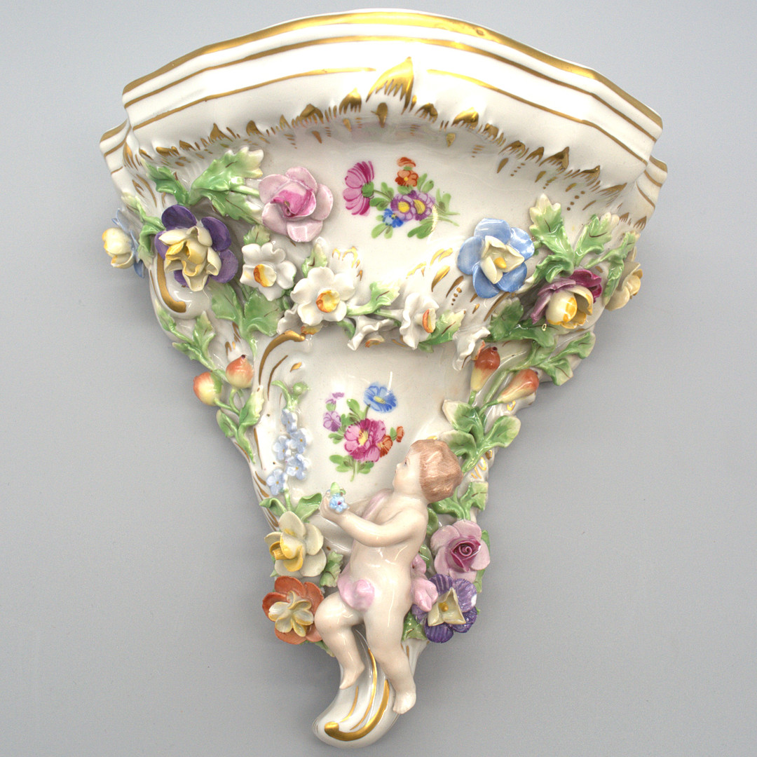 SÄCHSISCHE PORZELLAN-MANUFAKTUR DRESDEN POTSCHAPPEL. WANDKONSOLE MIT PUTTO, PLASTISCHEN BLÜTENBESATZ UND GOLD DETAILS. FRÜHES 20. JAHRHUNDERT.