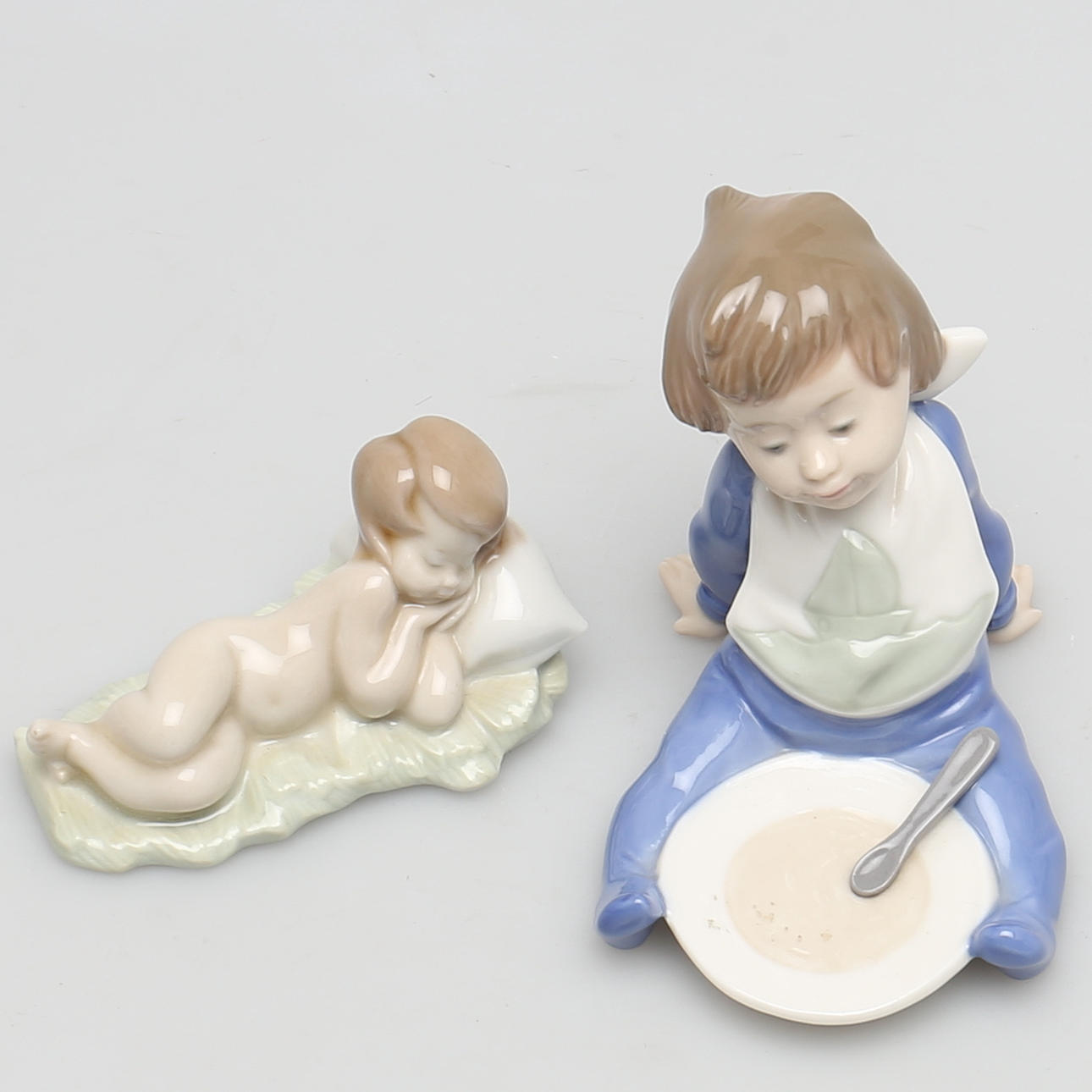 FIGURINER i porslin, 2 st, Pojke med grötfat och gosse mot huvudkudde och Nao.