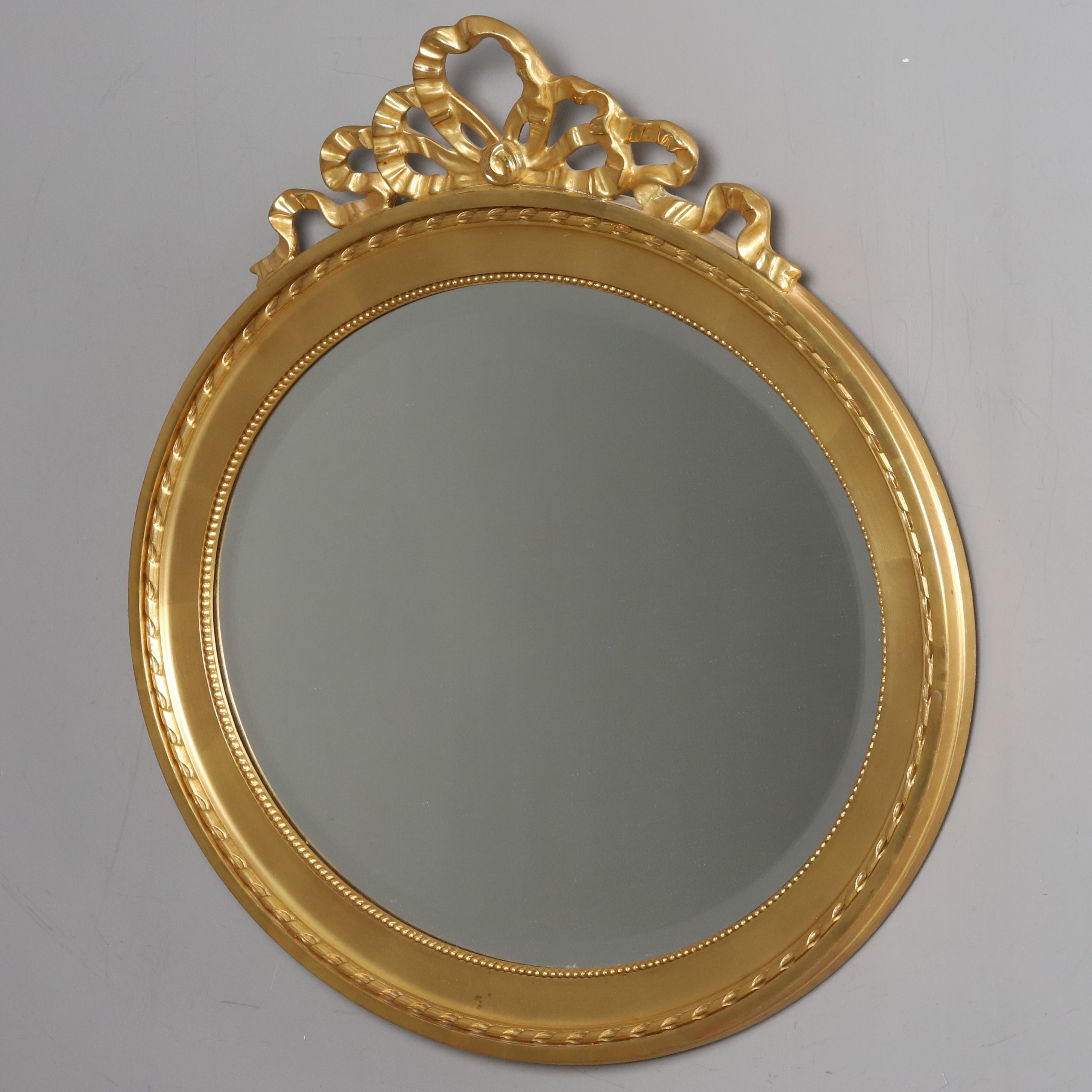 SPEGEL. Gustaviiansk-stil. Oval fasettslipad spegel, förgyllt trä med toppdekor av bandrosett. 1900-talets mitt.